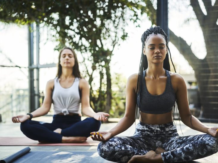 Young woman practicing lotus position with friend on porch. Multi-ethnic females are meditating in yoga class. They are in sports clothing.